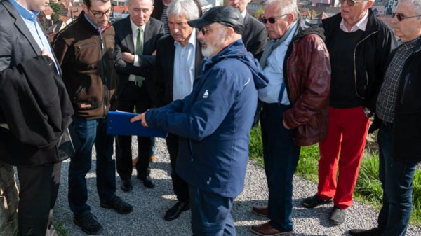 A group of twelve men, dressed in casual and business attire, gathers around a speaker who is gesturing with hands while holding a folder. They are outdoors, with a view of a cityscape in the background, surrounded by greenery and a stone wall.
