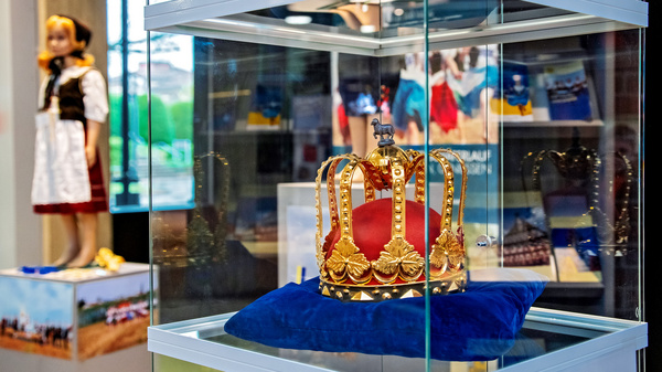 Eine gläserne Vitrine zeigt eine prächtige Krone mit roten und goldenen Verzierungen auf einem blauen Kissen. Im Hintergrund ist eine andere Vitrine mit einer Puppe in traditioneller Kleidung zu sehen.