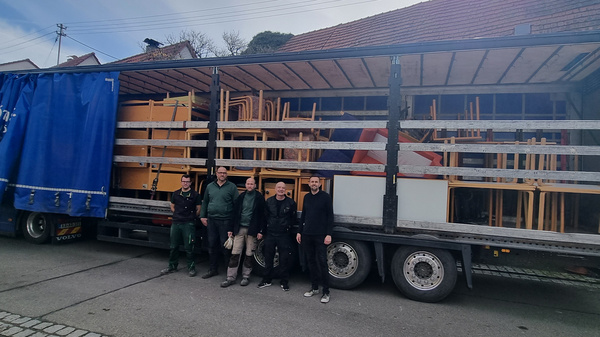 Ein Lkw steht auf einer Straße, beladen mit Tischen und Stühlen. Vor dem Lkw stehen fünf Personen in Arbeitskleidung. Im Hintergrund sind ein paar Gebäude und ein blauer Himmel zu sehen.