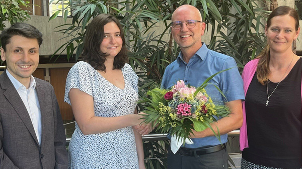 Vier Personen stehen in einem Innenraum, umgeben von Pflanzen. Eine Frau in einem weißen Kleid überreicht einem Mann mit Brille und blauem Hemd einen Blumenstrauß. Die anderen beiden Personen, ein Mann in einem dunklen Anzug und eine Frau in einem pinken Oberteil, stehen neben ihnen und lächeln.