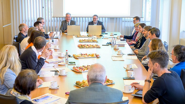 Ein Treffen in einem Konferenzraum, an einem großen Tisch, umgeben von Teilnehmern. Zwei Männer sitzen an der Kopfseite des Tisches und präsentieren. Auf dem Tisch sind Snacks und Getränke sichtbar. Die Atmosphäre wirkt fokussiert und kooperativ.