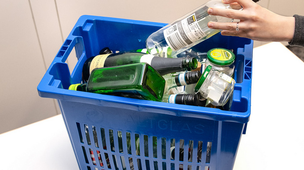 A blue plastic basket is filled with various empty liquor bottles, including green and clear glass containers. A hand reaches into the basket to hold a bottle, emphasizing the focus on recycling or disposal of glass bottles.