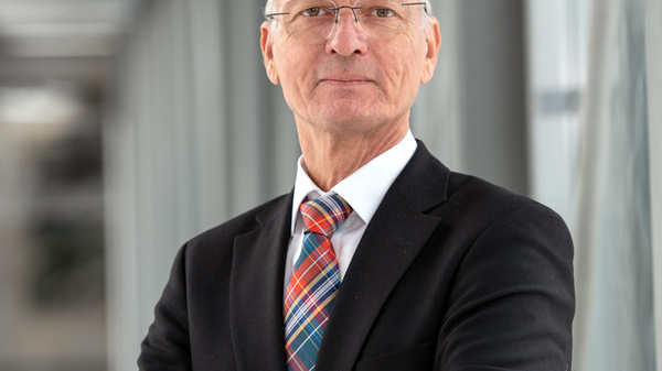 A confident older man in a black suit and colorful tie stands with his arms crossed. He is in a well-lit modern corridor, exuding professionalism and approachability.