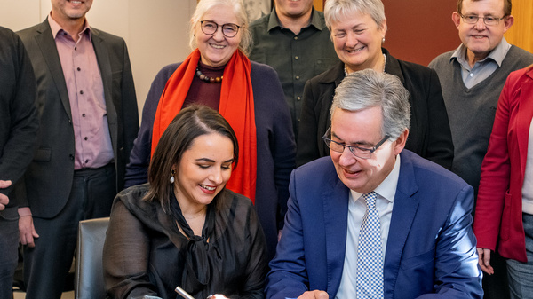 Eine Gruppe von Menschen steht um einen Tisch. Zwei Personen, eine Frau und ein Mann, sind im Vordergrund und unterschreiben ein Dokument. Die anderen schauen aufmerksam zu. Der Raum hat eine gemütliche Atmosphäre mit Holzpaneelen an den Wänden.