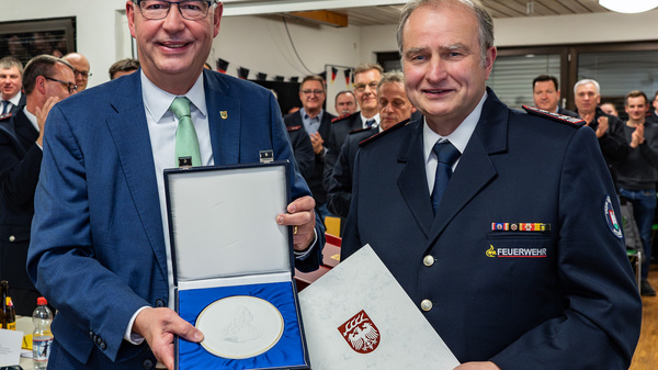 Zwei Männer stehen im Vordergrund und halten eine Auszeichnung in einer Schatulle. Der Mann links trägt einen Anzug und Brille, während der Mann rechts eine Feuerwehruniform trägt. Im Hintergrund ist eine Gruppe von applaudierenden Menschen zu sehen, die eine feierliche Stimmung vermittelt.