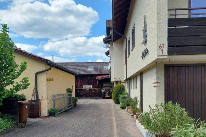 Eine Auffahrt zu einem Gebäude mit zwei Fenstern. An der linken Seite sind Pflanzen und ein Zaun sichtbar. Im Hintergrund sind weitere Gebäude und ein klarer Himmel mit Wolken zu erkennen. (Text KI generiert)