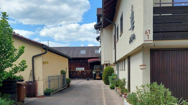 Eine Auffahrt zu einem Gebäude mit zwei Fenstern. An der linken Seite sind Pflanzen und ein Zaun sichtbar. Im Hintergrund sind weitere Gebäude und ein klarer Himmel mit Wolken zu erkennen. (Text KI generiert)
