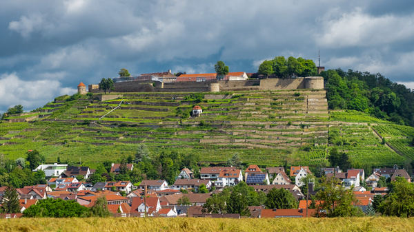 Une colline avec des vignes en terrasses surmontée par une forteresse. En bas, un village composé de maisons blanches et colorées. Le ciel est nuageux, ce qui ajoute une atmosphère de tranquillité au paysage.