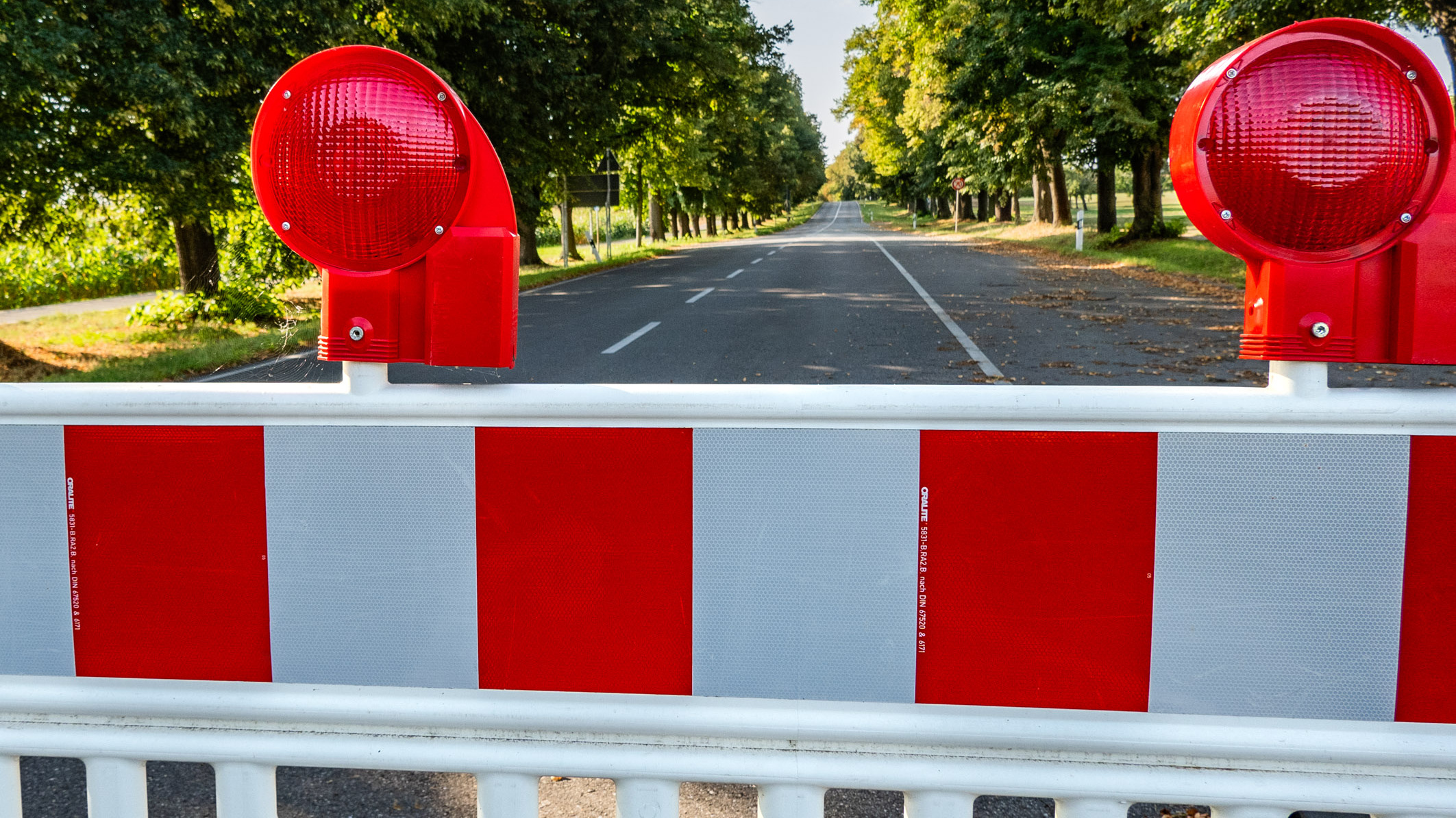 A red and white barrier blocks a deserted road flanked by green trees. Two red traffic lights indicate the closure of the road ahead, signaling to drivers that passage is restricted.