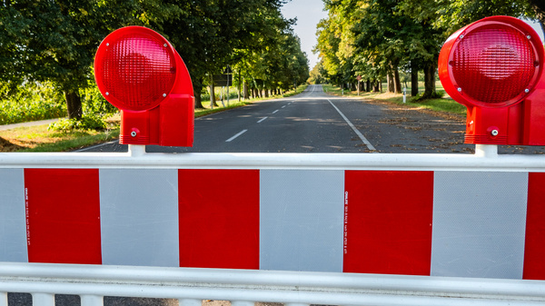 A red and white barrier blocks a deserted road flanked by green trees. Two red traffic lights indicate the closure of the road ahead, signaling to drivers that passage is restricted.