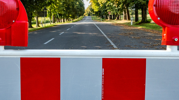 Eine Straßenbarriere mit roten Blinklichtern und weißen sowie roten Abschnitten ist im Vordergrund zu sehen. Im Hintergrund erstreckt sich eine leere Straße, die von Bäumen gesäumt ist, die im Sonnenlicht stehen. Die Barriere blockiert den Zugang zur Straße.