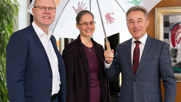 Three professionals stand together under a large umbrella decorated with colorful handprints. They are smiling and dressed in formal attire, with a background featuring a window and artwork on the wall. The image conveys teamwork and a positive collaboration spirit.