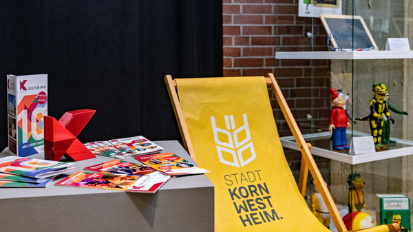 Ein gelber Liegestuhl mit dem Aufdruck „STADT KORNWESTHEIM“ steht vor einer Mauer. Darauf liegen bunte Broschüren und ein rotes Origami. Im Hintergrund sind verschiedene Ausstellungsobjekte zu sehen, die das Stadtleben und Veranstaltungen darstellen.