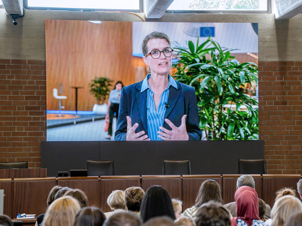 Eine Frau mit Brille steht vor einer großen Bildschirmpräsentation, spricht und gestikuliert. Im Hintergrund sind Pflanzen zu sehen und Teilnehmer einer Veranstaltung sind in der unteren Bildhälfte versammelt. Das Bild vermittelt eine kommunikative und aufmerksame Atmosphäre.