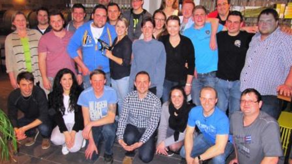 A large group of diverse individuals is gathered together, posing for a photo indoors. They are arranged in a semi-circle, smiling, with some holding a trophy, suggesting a celebratory occasion. The background features shelves lined with bottles.