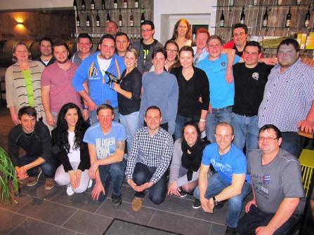 A large group of diverse individuals is gathered together, posing for a photo indoors. They are arranged in a semi-circle, smiling, with some holding a trophy, suggesting a celebratory occasion. The background features shelves lined with bottles.