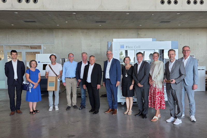 Eine Gruppe von zwölf Personen steht in einem modernen Innenraum. Sie lächeln und posieren gemeinsam für das Bild. Im Hintergrund sind verschiedene Ausstellungsstände sichtbar, darunter ein "Flex Cell"-Stand. Die Stimmung wirkt freundlich und einladend.