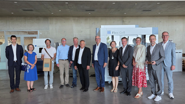 Eine Gruppe von zwölf Personen steht in einem modernen Innenraum. Sie lächeln und posieren gemeinsam für das Bild. Im Hintergrund sind verschiedene Ausstellungsstände sichtbar, darunter ein "Flex Cell"-Stand. Die Stimmung wirkt freundlich und einladend. (Text KI generiert)