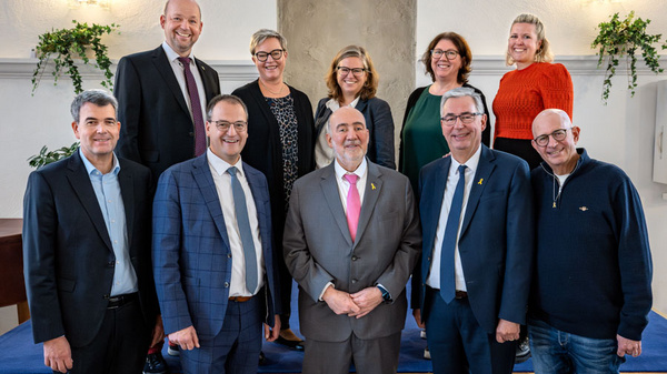 Eine Gruppe von neun Personen steht auf einer kleinen Bühne. Sie lächeln und präsentieren sich freundlich. Im Hintergrund sind Pflanzen und eine dekorative Wand. Die Personen tragen unterschiedliche formelle Kleidung und scheinen in einer festlichen Atmosphäre zusammenzukommen.
