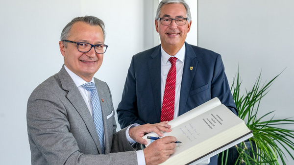 Two men are standing beside a large open book on a stand. One man is smiling and holding a pen, poised to write, while the other is looking at the camera. Both are dressed in formal attire, and there is a potted plant in the background.