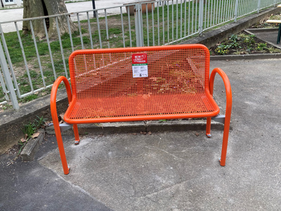 Panchina arancione in metallo situata in un'area verde. È priva di occupanti e presenta un cartello sopra lo schienale. Il pavimento è in cemento, con alcune foglie visibili attorno.