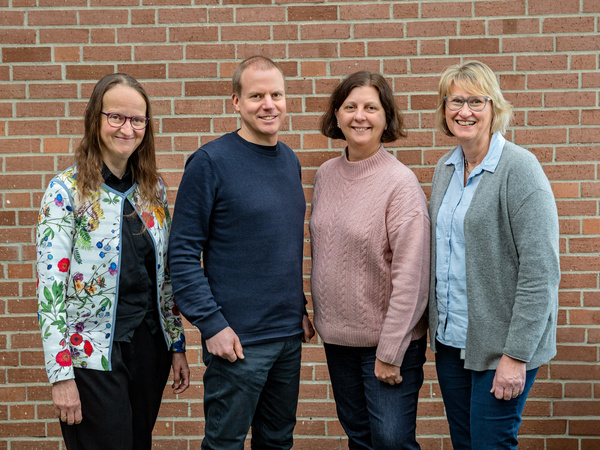 Vier Personen stehen in einer Reihe vor einer Backsteinwand. Sie lächeln und tragen legere Kleidung. Die Frauen haben unterschiedliche Frisuren und Outfits, der Mann ist in einem dunkelblauen Pullover gekleidet. Die Szene wirkt freundlich und einladend.
