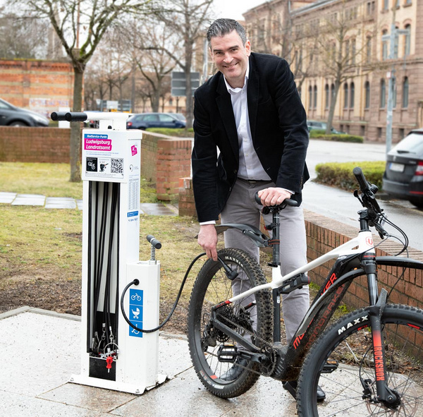 Ein Mann steht neben einer Fahrradreparaturstation. Er hält ein Mountainbike, während er an einer der mit Schläuchen ausgestatteten Station arbeitet. Im Hintergrund sind Bäume und Gebäude sichtbar.