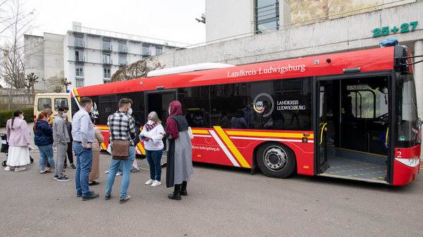 Eine Gruppe von Menschen wartet, um in einen roten Bus mit der Aufschrift "Landkreis Ludwigburg" einzusteigen. Der Bus steht an einer Haltestelle, während im Hintergrund ein Gebäude sichtbar ist. Die Wartenden sind verschieden gekleidet und mehrere scheinen Gespräche zu führen.