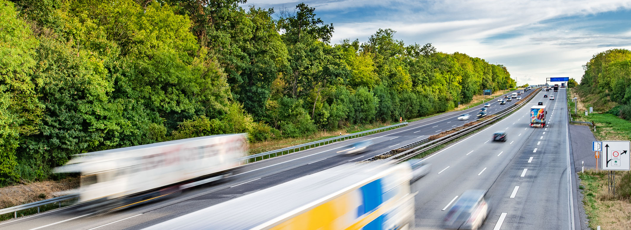 Header Bild: Auf der Bildmitte ist eine stark befahrene Autobahn zu sehen, auf der verschiedene Lastwagen und PKWs in beide Richtungen fahren. Im Hintergrund sind dicht bewaldete Gebiete und ein blauer Himmel mit Wolken sichtbar. Die Fahrzeuge scheinen sich schnell zu bewegen.