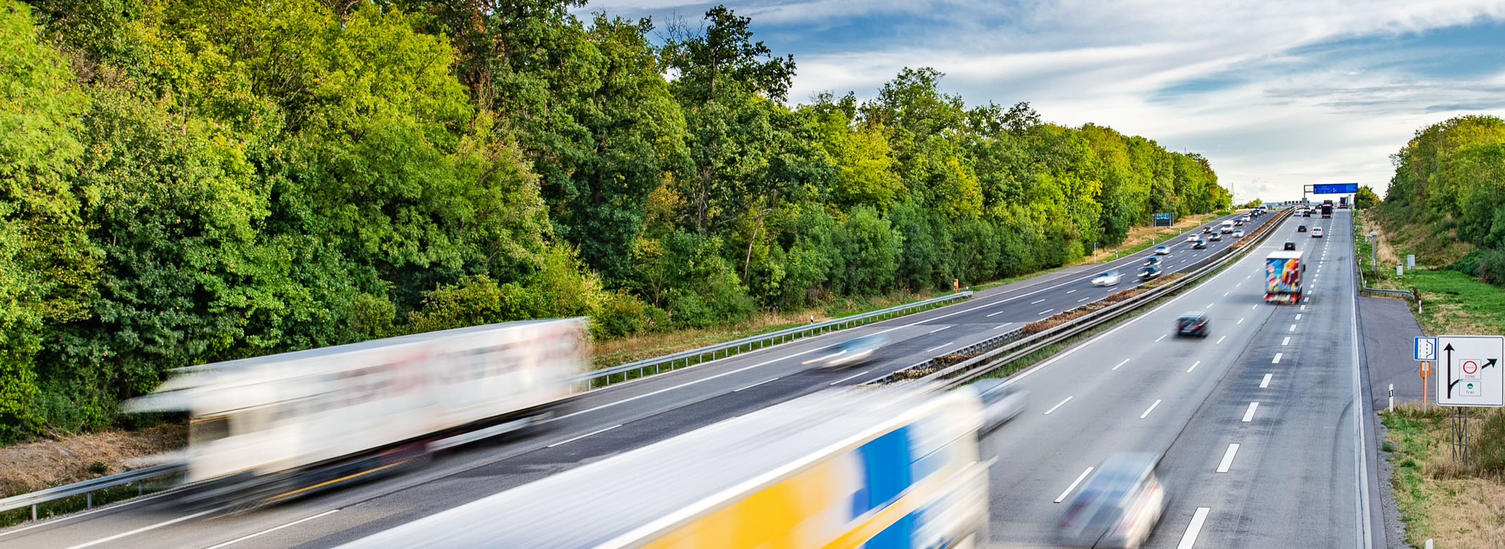 Header Bild: Auf der Bildmitte ist eine stark befahrene Autobahn zu sehen, auf der verschiedene Lastwagen und PKWs in beide Richtungen fahren. Im Hintergrund sind dicht bewaldete Gebiete und ein blauer Himmel mit Wolken sichtbar. Die Fahrzeuge scheinen sich schnell zu bewegen.