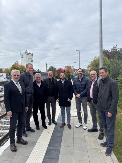 Eine Gruppe von neun Männern steht auf einem Bahnsteig. Sie tragen verschiedene Business-Outfits und lächeln in die Kamera. Im Hintergrund sind Bäume mit buntem Laub und ein Gebäude sichtbar. Der Himmel ist bewölkt.
