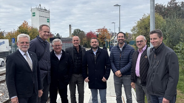 Eine Gruppe von neun Männern steht auf einem Bahnsteig. Sie tragen verschiedene Business-Outfits und lächeln in die Kamera. Im Hintergrund sind Bäume mit buntem Laub und ein Gebäude sichtbar. Der Himmel ist bewölkt.
