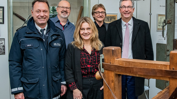 Eine Gruppe von fünf Personen steht in einem Museum. Im Vordergrund ist eine alte Holzmaschine zu sehen. Die Personen lächeln, sie tragen unterschiedliche Kleidung, darunter eine Uniform und eine schwarze Jacke. Der Hintergrund zeigt Wandtafeln mit historischen Bildern.