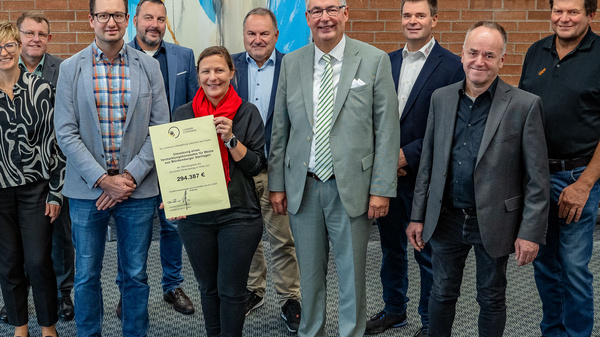 Eine Gruppe von zwölf Personen steht in einem Raum vor einer gemusterten Wand. Sie halten ein Zertifikat in der Mitte. Die Gruppe besteht aus Männern und Frauen unterschiedlichen Alters, die lächeln und in formeller Kleidung gekleidet sind.