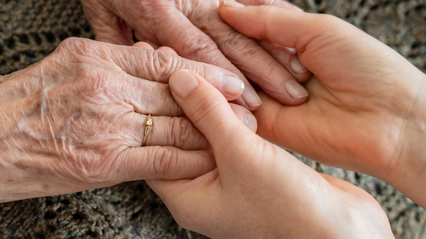 Zwei Hände halten sich sanft, eine ältere Hand mit sichtbaren Adern und einer Ring, die andere jünger und zart. Diese Berührung symbolisiert Nähe und Unterstützung zwischen den Generationen, auf einem strukturierten Textiluntergrund.