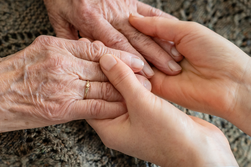 Zwei Hände halten sich sanft, eine ältere Hand mit sichtbaren Adern und einer Ring, die andere jünger und zart. Diese Berührung symbolisiert Nähe und Unterstützung zwischen den Generationen, auf einem strukturierten Textiluntergrund.