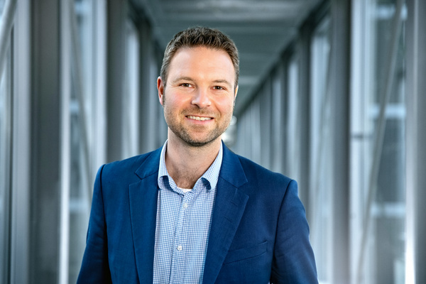 Ein lächelnder Mann mit Kurzhaarschnitt trägt einen blauen Anzug und ein karriertes Hemd. Er steht in einem modernen, hellen Gang mit großen Fenstern im Hintergrund, das eine professionelle und freundliche Atmosphäre vermittelt.