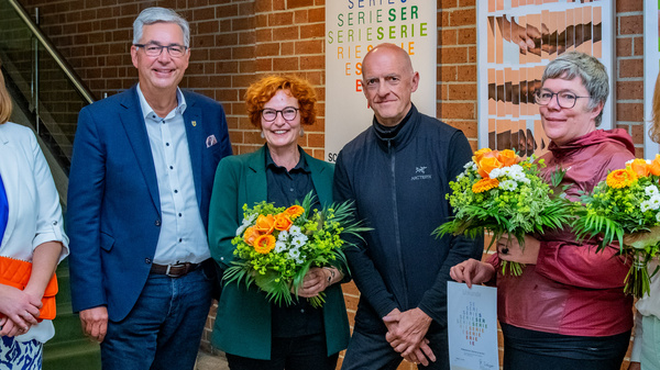 Eine Gruppe von sechs Personen steht vor einer Wand mit Kunstwerken. Alle halten Blumensträuße. Zwei Personen tragen formelle Kleidung, während die anderen lässiger gekleidet sind. Im Hintergrund ist eine Informationswand zur Ausstellung zu sehen. Die Stimmung ist festlich und freundlich.