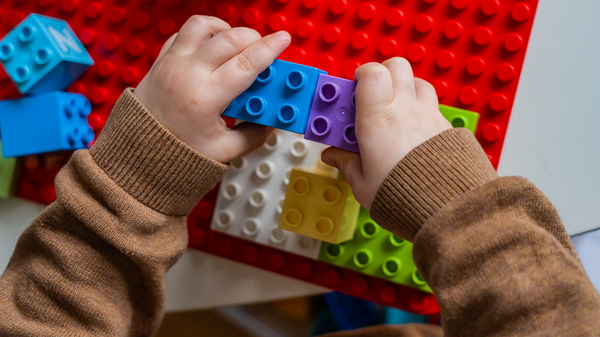 Ein Kind mit Händen in einem braunen Pullover baut mit bunten Lego-Steinen. Es hält einen lila und einen blauen Stein, während es versucht, sie zusammenzufügen. Im Hintergrund sind weitere Lego-Steine und eine rote Grundplatte sichtbar. (Text KI generiert)