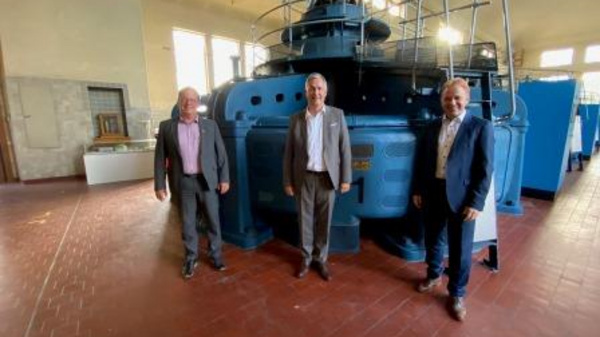 Three men stand in front of a large, blue industrial machine in a spacious, well-lit room. The machine has a cylindrical shape and features multiple levels and railings. The men are dressed in formal attire, smiling at the camera.