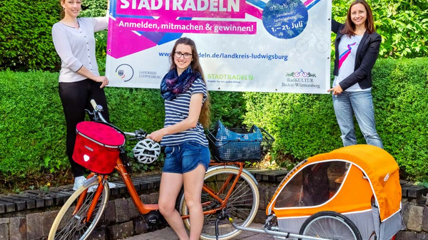 Three people stand outdoors next to a bicycle and a trailer. They are holding a large banner promoting "Stadtradeln," an initiative encouraging cycling. The banner includes details about registration and participation. Lush greenery serves as the background for this promotional event.