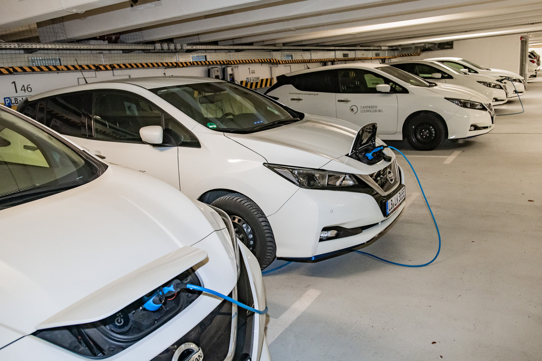 Weißer Elektroautos, die in einer Parkgarage aufgeladen werden. Ein Auto ist mit einem Kabel an einer Ladestation verbunden, während andere Fahrzeuge im Hintergrund stehen. Der Boden ist hell und sauber, die Garage wirkt modern. (Text KI generiert)