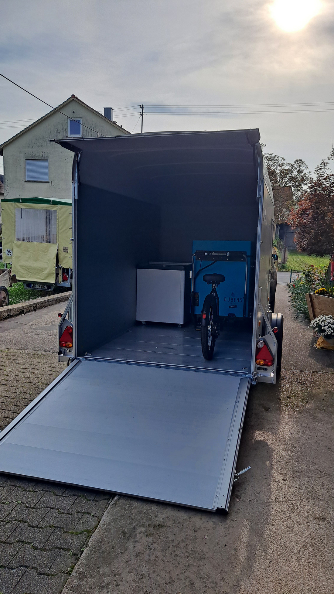 Ein offener Anhänger steht auf einer gepflasterten Fläche. Im Inneren sind ein Fahrrad und ein Kühlschrank sichtbar. Der Anhänger hat eine herunterklappbare Rampe, und im Hintergrund ist ein Wohnhaus zu erkennen. Die Sonne scheint und der Himmel ist teilweise bewölkt.