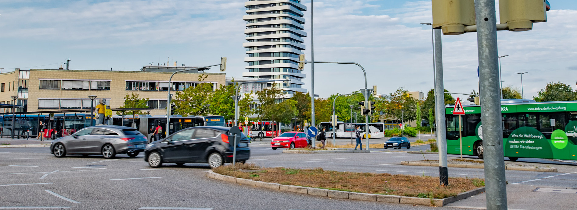 Header Bild: Eine städtische Szene mit Verkehrszeichen für Radfahrer und Fußgänger. Im Hintergrund ist ein modernes Hochhaus zu sehen, umgeben von Geschäftsgebäuden und einer Bushaltestelle. Mehrere Autos sind auf der Straße unterwegs. Der Himmel ist teilweise bewölkt.