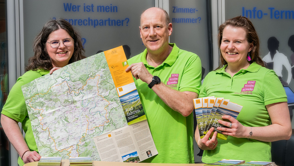 Three people stand behind a display table. They are dressed in bright green shirts and are smiling. One person holds a large map, while another holds brochures. The table features brochures and a scenic image of vineyards. The background includes text in German.