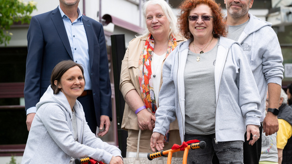 Sechs Personen stehen zusammen im Freien. Eine Frau sitzt auf einem orangefarbenen Kinderfahrrad. Sie lächeln und wirken glücklich. Es gibt eine grüne Umgebung im Hintergrund, einschließlich Bäume und Gebäude.