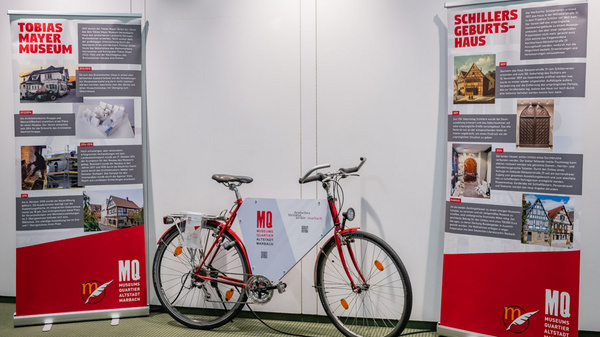 Das Bild zeigt ein Fahrrad in der Mitte, umgeben von zwei Informationsständen. Die Stände präsentieren Informationen über das Tobias Mayer Museum und Schillers Geburtshaus, mit textlichen Details und Bildern. Die Szenerie ist hell und modern gestaltet.