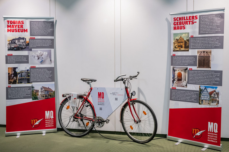 Das Bild zeigt ein Fahrrad in der Mitte, umgeben von zwei Informationsständen. Die Stände präsentieren Informationen über das Tobias Mayer Museum und Schillers Geburtshaus, mit textlichen Details und Bildern. Die Szenerie ist hell und modern gestaltet.