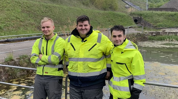 Drei Personen in hellen Arbeitsjacken stehen lächelnd auf einer Brücke über einem Gewässer. Im Hintergrund sind Grünflächen und eine Anhöhe zu sehen. Es handelt sich um eine Arbeitsumgebung, wahrscheinlich im Bereich Umweltschutz oder Ingenieurwesen.