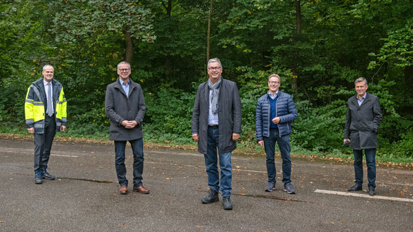 Five men stand on a road in a wooded area. They are dressed in various coats and jackets, with two men in lighter outerwear. The scene appears to depict a professional or formal gathering outdoors. Trees and greenery are visible in the background.